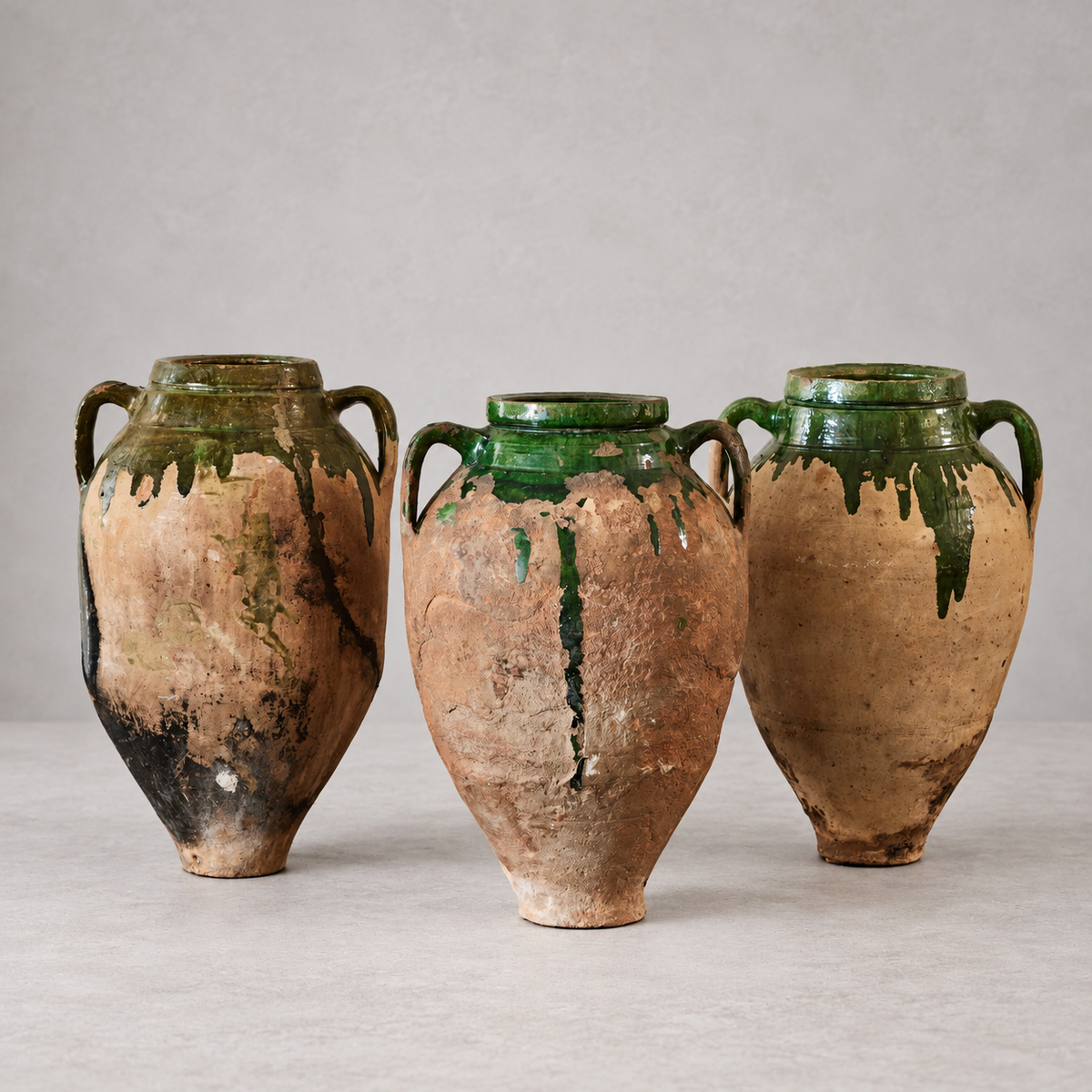 Three antique Bulgarian terracotta olive oil pots with twin handles, weathered surfaces, and green glaze dripping from their shoulders, spread apart