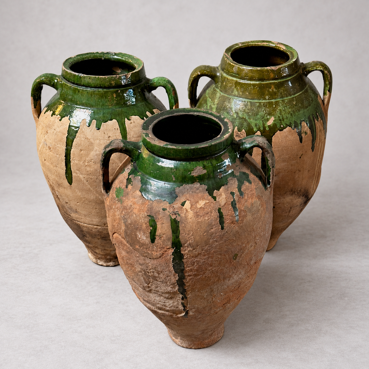 Three rustic ceramic amphora-style vases with two handles, featuring a distressed terracotta-brown body and glossy dark green glaze dripping down the sides, set against a neutral light gray background.