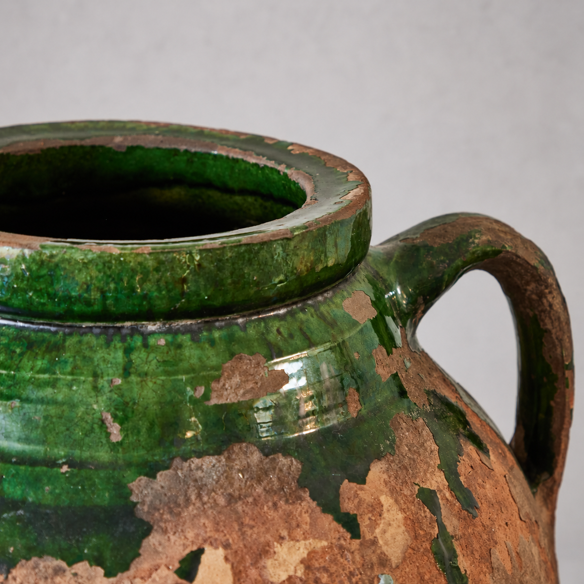 Close-up of a terracotta olive oil pot’s worn green-glazed rim and curved handle, with chipped glaze exposing the clay beneath.