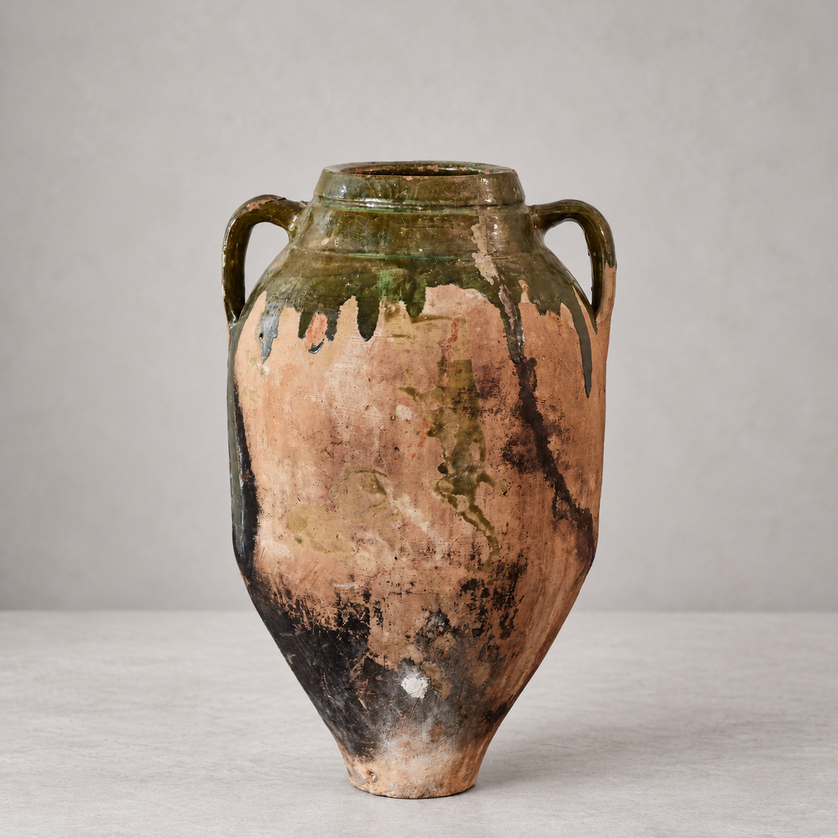 An antique ceramic vase with two handles, featuring a distressed terracotta body with dark brown and green glaze drips, set against a neutral gray background.